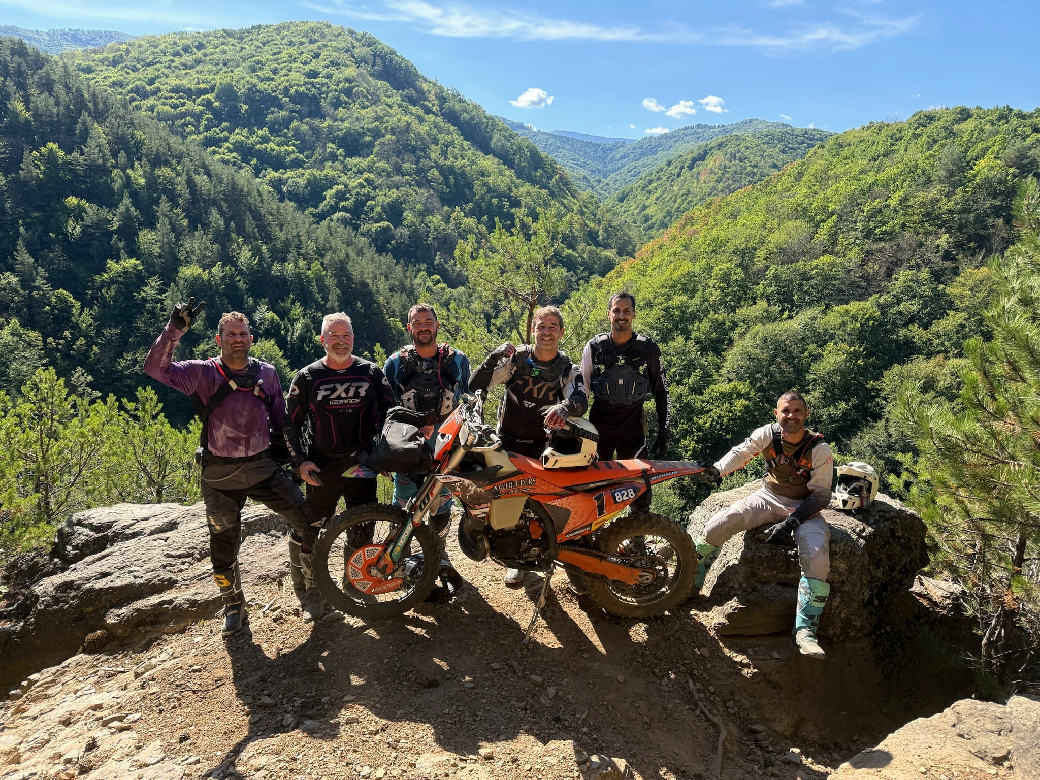 Seven riders on a rock outcrop above a forested valley, one with his arm raised, an orange KTM in front of them