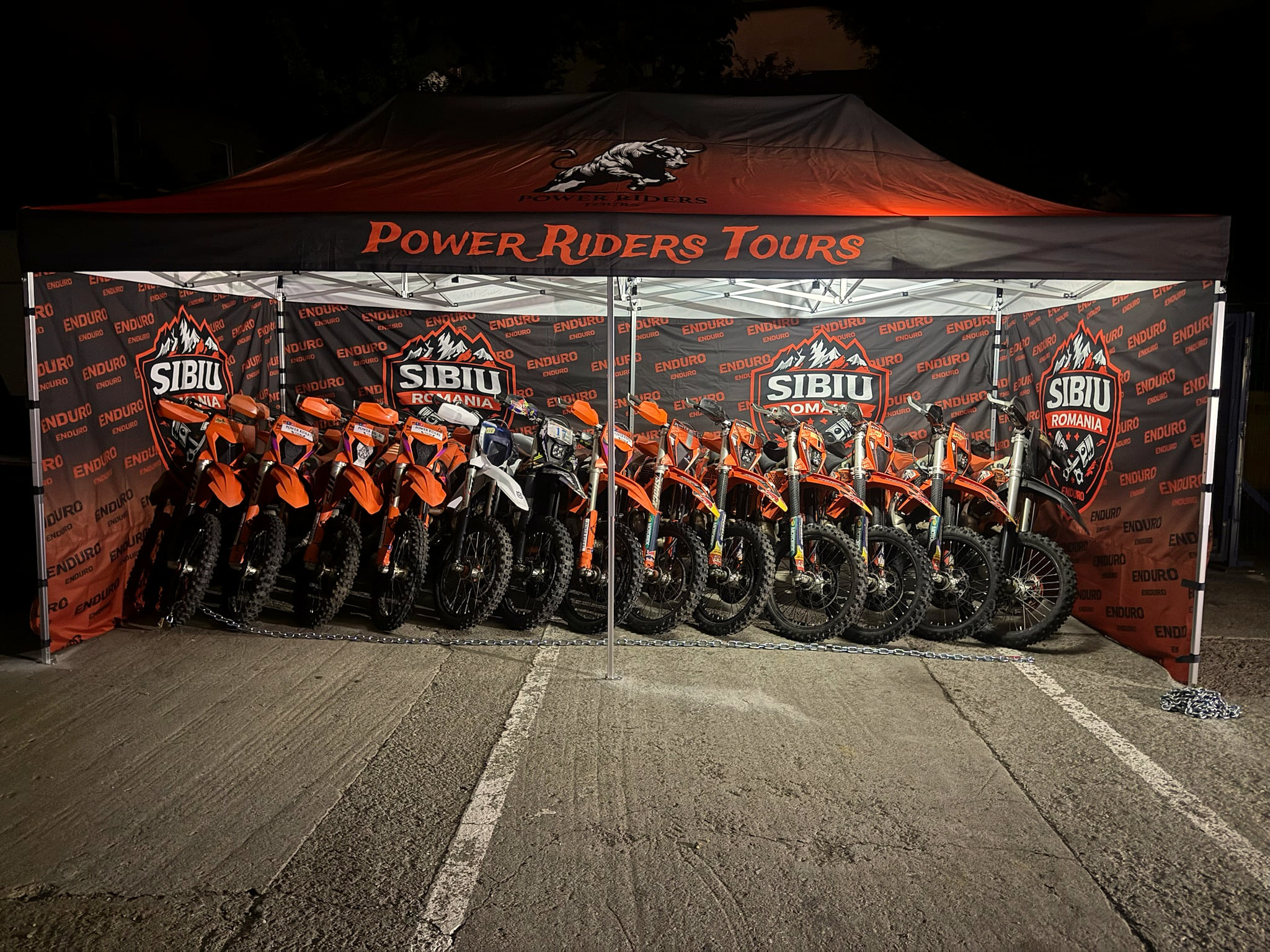 Orange KTM enduro bikes lined up under the Power Riders Tours tent, Sibiu banners on its walls.