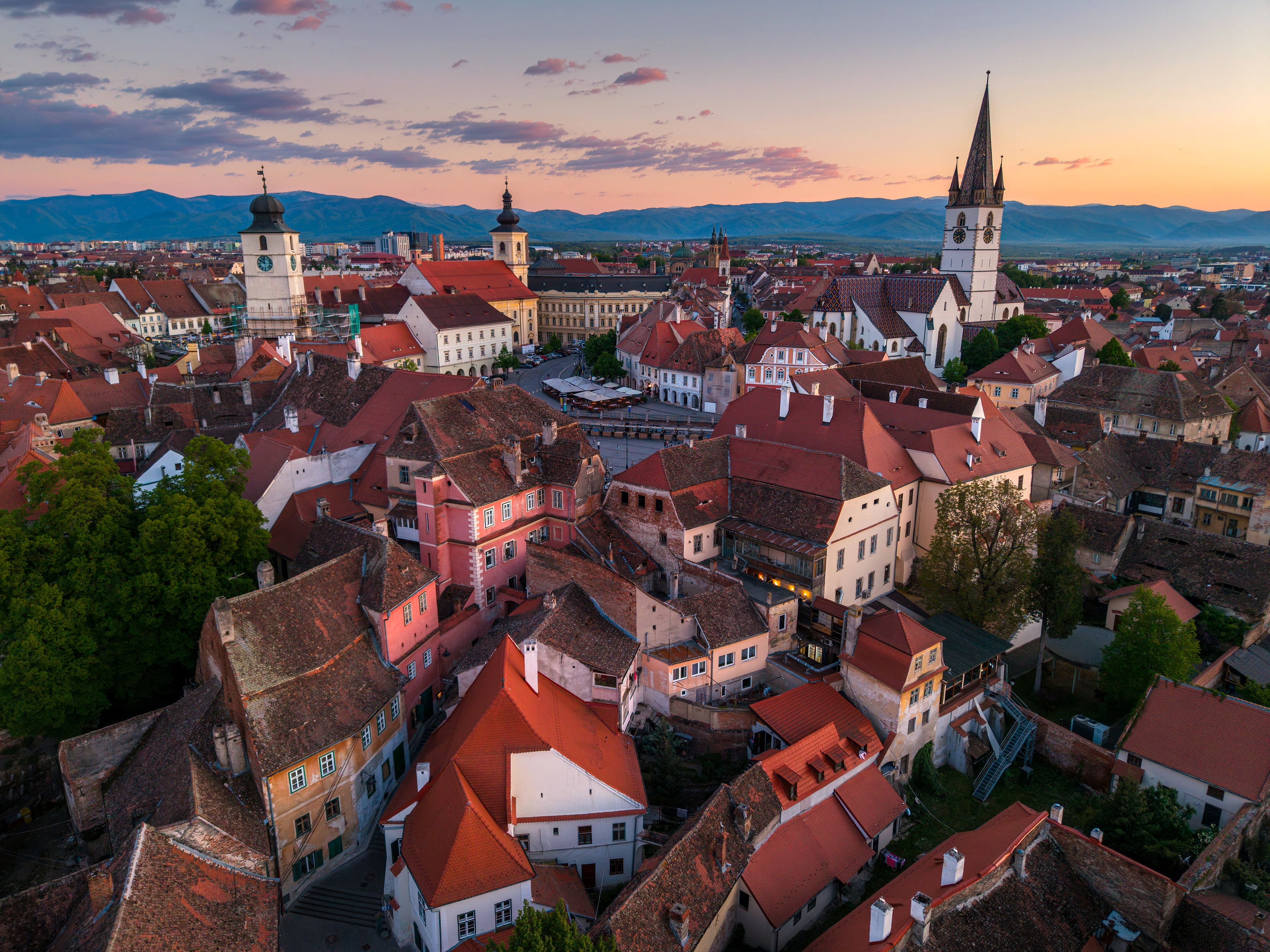 Sibiu's old town from the air at dusk: red roofs, old towers and the Lutheran Cathedral, with mountains behind.
