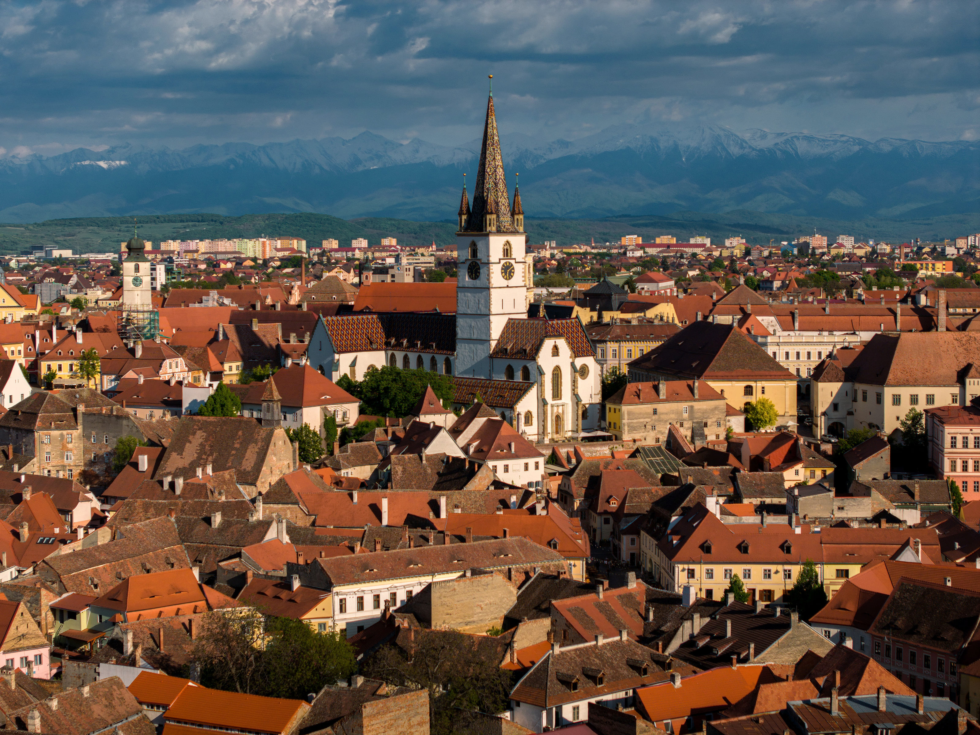 Red roofs of Sibiu's old town around the Lutheran Cathedral's spire, snow-capped mountains on the horizon.